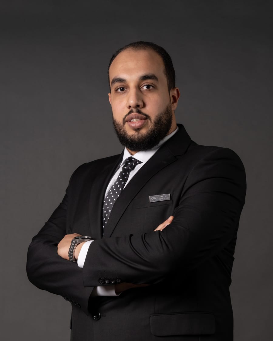 Hesham Salah in a dark suit, arms folded, against a charcoal studio background